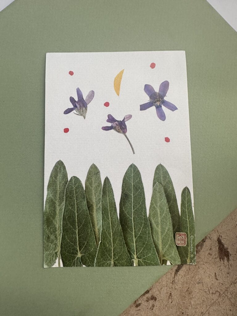 A postcard with 8 leaves on the bottom half, 3 lavender flowers on the top half with 5 red dots and a half moon. The postcard has white background.