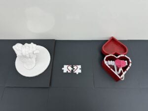 A mold of 2 hands holding each other, 2 puzzle pieces each with half of a heart, and a red wooden heart-shaped box with a pop up heart inside