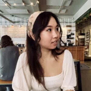 The artist, Joanne Kwon, wearing a white top and floral bandana, looking off to the side with a gentle smile.