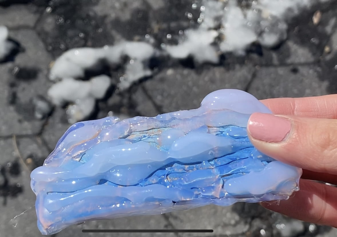 On the right, I hold the tactile graphic in my hands in front of ice on the concrete ground. The graphic is an inch tall and two inches wide. It has a blue oil pastel base and layers of hot glue that crest over the top edge of the cardboard and catch the sun.