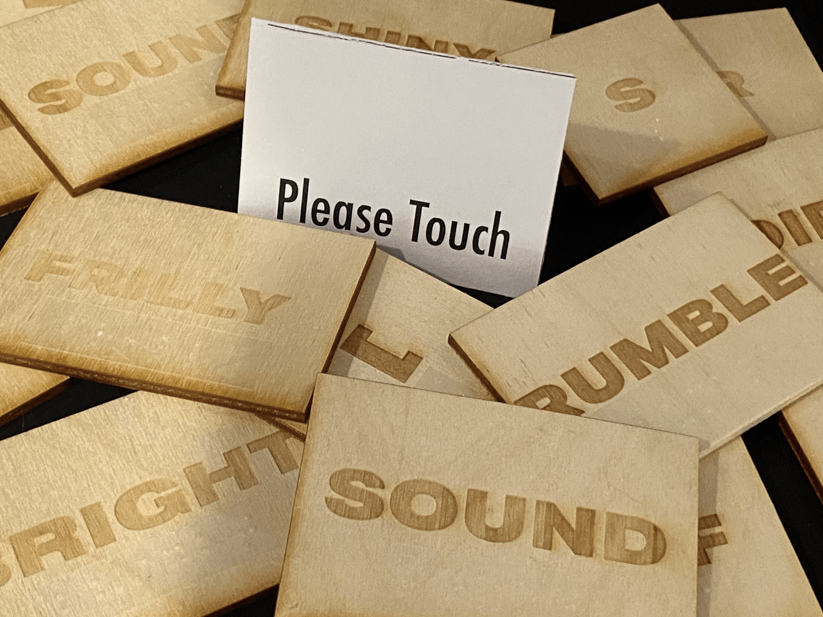 a pile of wooden tiles on a black surface. a folded white card reads "please touch"