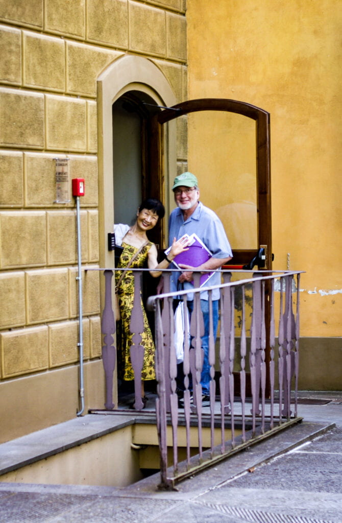 Ikuko stands in a doorway with a colleague and waves cheerfully.