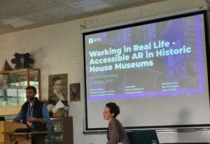 Craig stands at a podium wearing a black vest and blue flannel. Seated to the right is Rosanna, who wears a brown cardigan. Behind them is a purple powerpoint slide with white text reading "Working in Real Life - Accessible AR in Historic House Museums". 