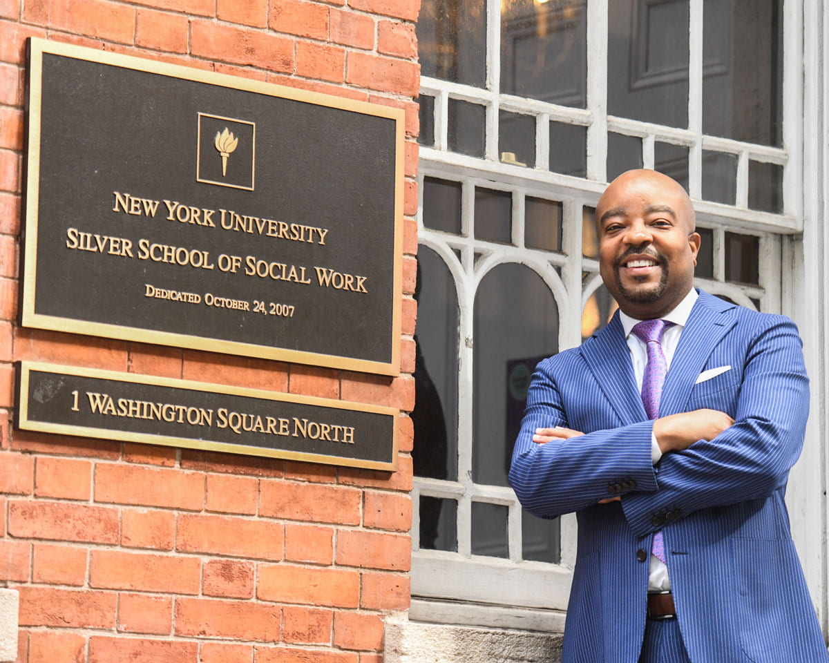 Dean Lindsey stands at the entrance of 1 Washington Square North next to the NYU Silver plaque