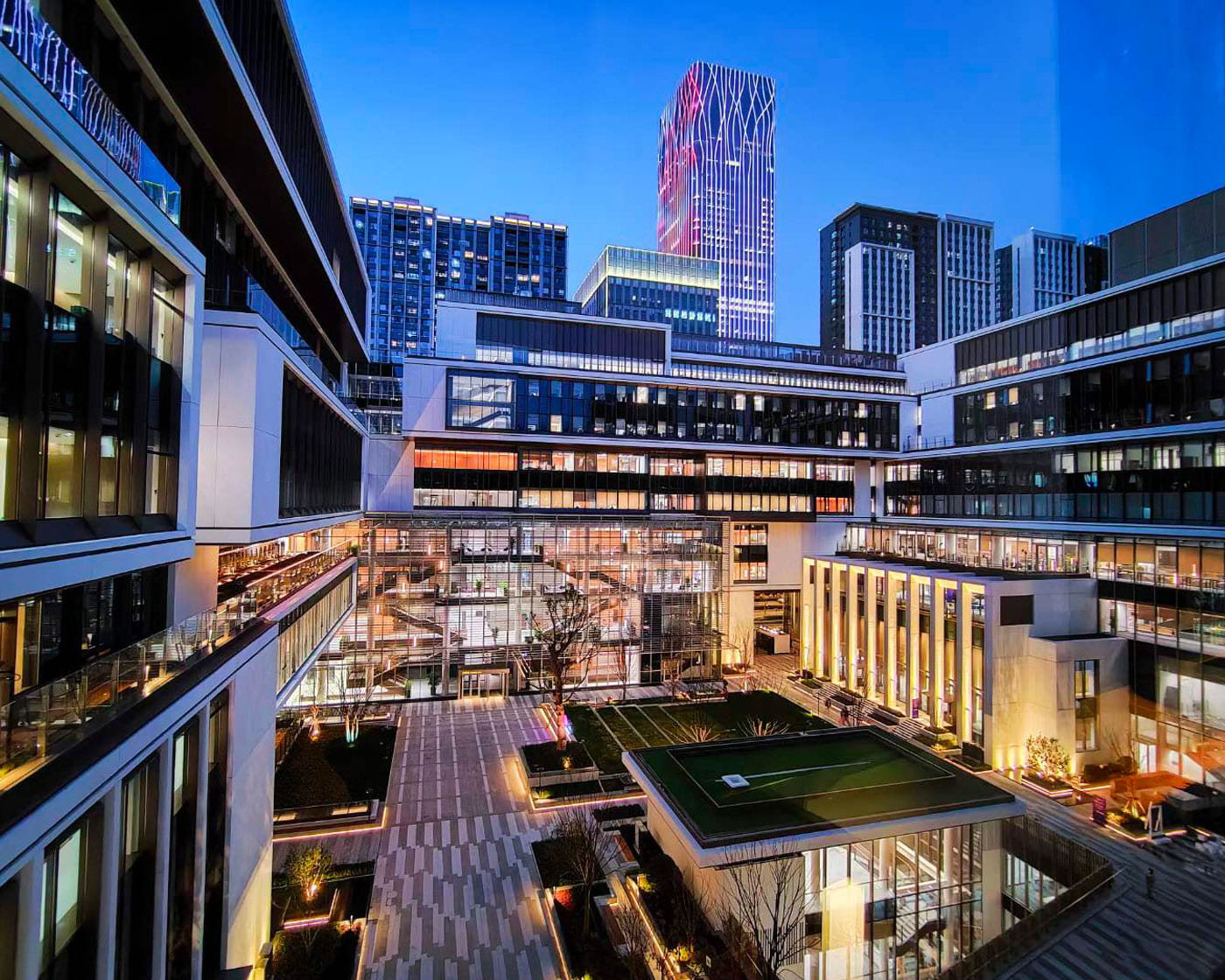 Nighttime view of NYU Shanghai's Bund campus buildings with warmly lit floor to ceiling windows and skyscrapers in the background