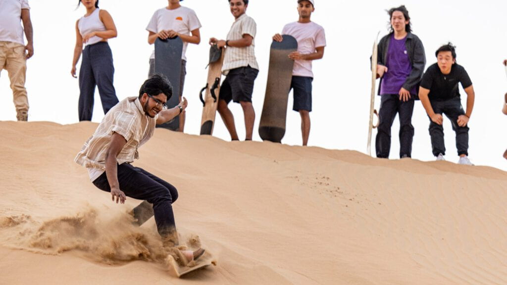 A student surfing down a hill of sand as other students wait for their turn.