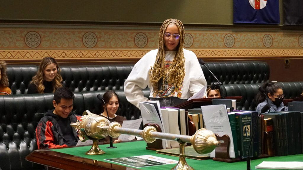 A student role playing at parliament, while other students are sitting in rows behind them.