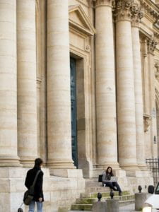 A young woman on a laptop sits on the steps to the Sorbonne, a building with large columns.