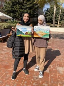 Two women in head scarves hold up paintings