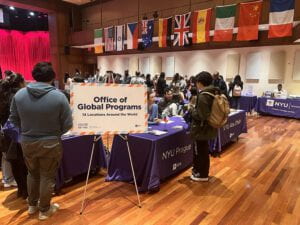 A sign reads, "Office of Global Programs. 14 Locations Around the World." and stands near students standing at seated at tables.