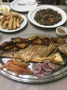 A plated meal of fish, plantains, onion, sauteed beans and vegetables