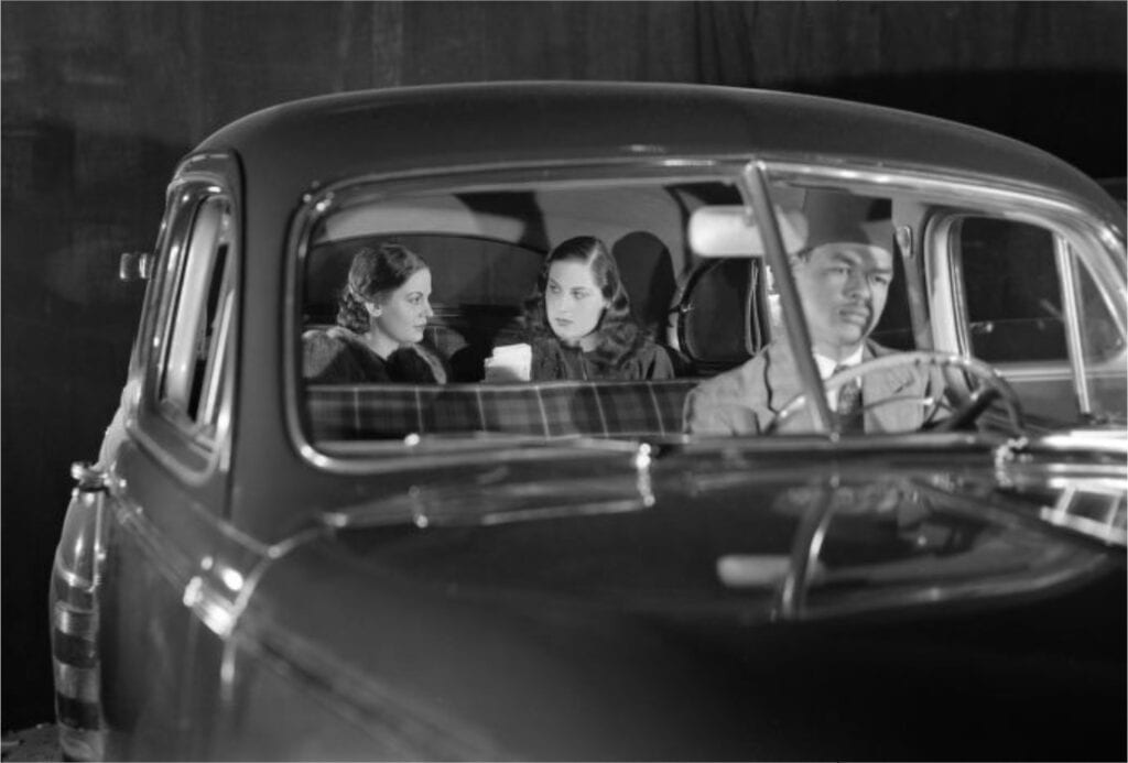 A black and white image of two woman in the back of a car with a male drive in the front.