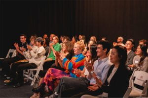 The summit audience, seated, clapping and smiling.