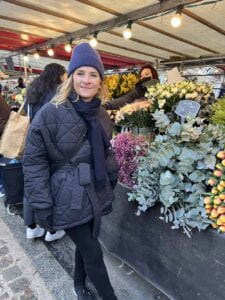 Martina Faltova in a coat and hat stands in front of a flower stand