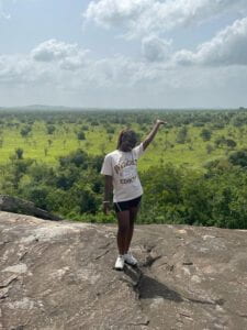 Julia Antwi-Boasiako poses from rocky terrain in front of a green field of trees with hills in the background