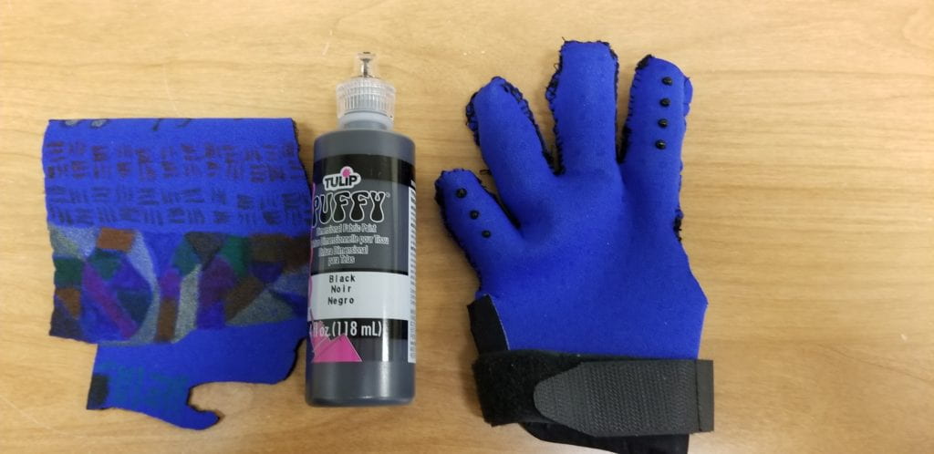 THE GLOVE IS ON THE RIGHT AND A TUBE OF PUFFY PAINT IS IN THE MIDDLE. THE GLOVE HAS PUFFY PAINT SPOTS ADDED TO THE INDEX FINGER AND THE LITTLE FINGER OF THE VOLAR ASPECT OF THE GLOVE. ON THE LEFT IS A PRACTICE PIECE OF MATERIAL WITH FABRIC PAINT