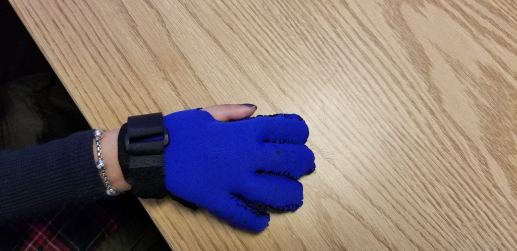 A D-RING STRAP SEWN TO THE BASE OF THE GLOVE AND PATIENT IS PLACING THE GLOVE ON THE TABLE WHILE ON THE HAND