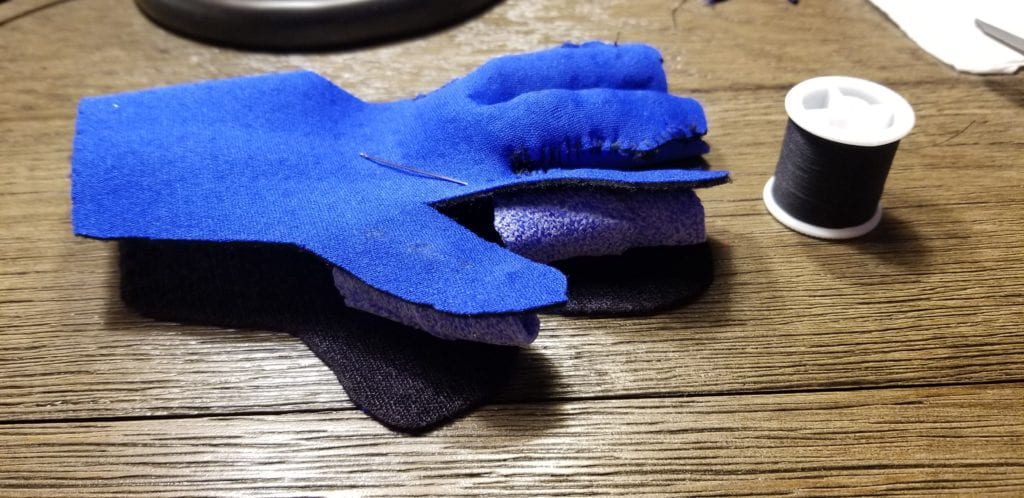 SIDE VIEW OF THE GLOVE HALF-SEWN WITH THE ORFICAST INSERTS AND A BLACK SPOOL OF THREAD BESIDE THE GLOVE. THREADING NEEDLE ON TOP OF THE GLOVE.