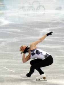 figure skater with black leggings and a white and black top wearing black gloves and in a sit spin position on the ice