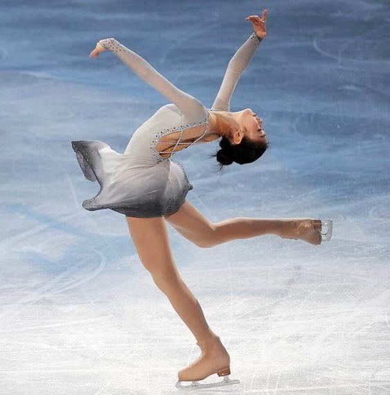 A female-presenting figure skater in a white and gray dress arches her back as she glides across the ice.