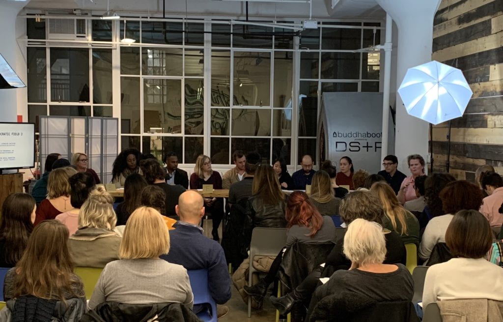 Actors sitting at a long table in the front of a large room reading policy statements for an audience.