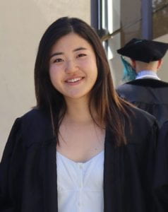 Mid-torso photo of Lydia smiling while wearing her black graduation gown and light blue dress underneath.