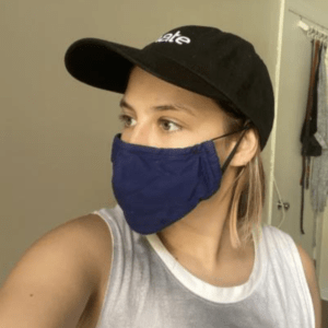 Portrait photo of student (white female with short blond hear) looking at the right side from the camera wearing the modified blue mask with the black cap with the mask elastic going above the ear.