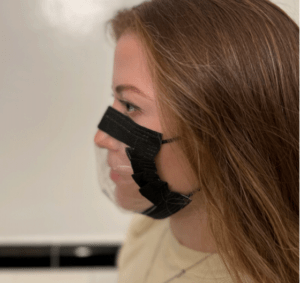 Student shown in profile view, facing to the left. The mask is adapted from a black medical mask, with the center of the mask removed and replaced with a clear, plas>c rectangle. The plas>c por>on of the mask is sewn to the black perimeter using black thread