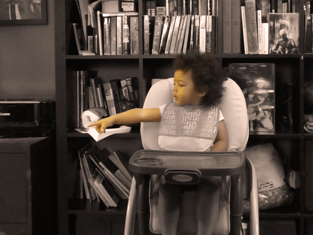 toddler is seated in a high-chair pointing at something in the distance