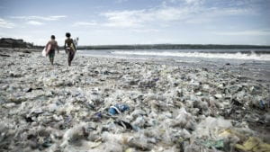 Two surfers walk on a beach completely littered with plastic waste