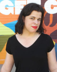 A woman with dark hair in a black t-shirt and red lipstick sits in front of a painted wall.