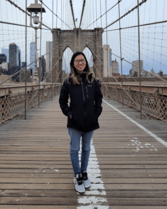 A young woman in jeans and a black jacket stands on a bridge.