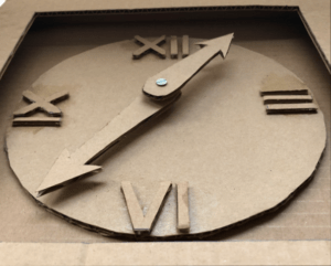 closeup of clock face made out of cardboard featuring hour and minute hands, and roman numerals for 3, 6, 9, and 12. 