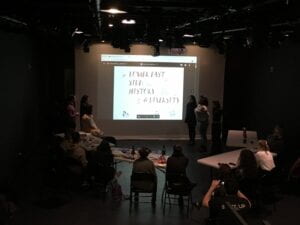 Image of students presenting in front of a large screen in a dark theater space