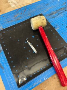 A black sheet of silicone rubber with holes, on top of a blue mat with a tan and red mallet and metal poker