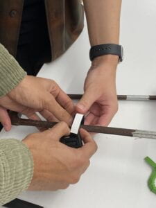 A member of the group holds a Fidlock slide magnet in place while raising the arm of a harness slightly over a table. Another member tries looping a velcro strap through the slot on the edge of the of the slide magnet.