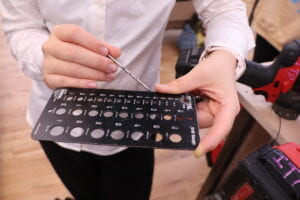 A woman measuring a drill bit with a metal sheet with multiple nail diameters.