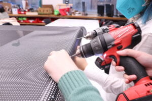 Two women drilling holes and fastening nails to trampoline mesh on top of a PVC frame