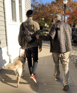 The image shows Mashi and Lukas walking with Beau, a guide dog at the Seeing Eye