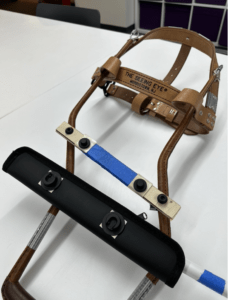 A thin piece of wood lies across the two legs of a harness, held in place by a plumbing clamp on either side tightened with a screw. Two Fidlock Snap plugs are attached along the wooden base. Their corresponding sockets are attached to the sheath, facing opposite directions to facilitate a twisting motion when detaching. The sheath is placed along the legs of the harness with the cane sticking out through its opening.