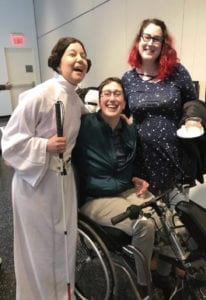 IMAGE DESCRIPTION: A photograph from the CDS- supported exhibition “Crip Imponderabilia” at NYU’s Gallatin Gallery, April 2019. Facing the camera are curator Bojana Coklyat, standing with a cane, dressed in a Princess Leia costume. To her right are disability activists, performers and educators Visiting Professor in Performance Studies Danielle Peers, seated in a wheelchair-cycle, and Dr. Lindsay Eales who stands behind.