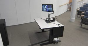 IMAGE DESCRIPTION: The photo shows an electrically powered adjustable height table set up for use in an NYU lecture hall. The set up includes a computer, wireless and wired keyboard, microphone, large computer monitor on an adjustable arm which also serves as the interface to an integrated Crestron media control system which is incorporated in the computer/monitor.