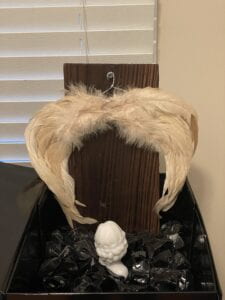 black box full of black rocks, a the top half of a woman made of white resin, a dark brown wooden board with wings hanging from it