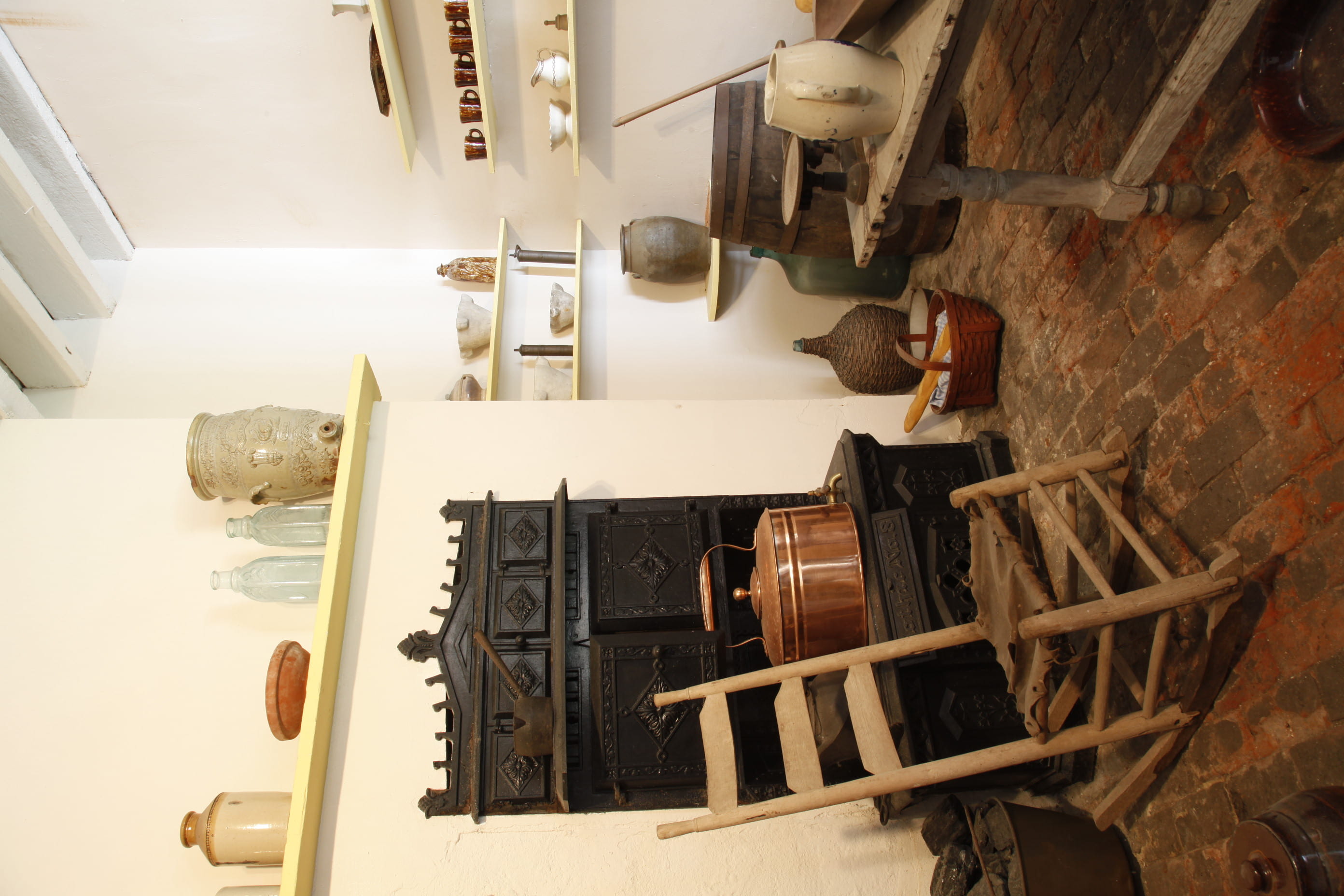 A picture of a historical room with a black stove, a wooden rocking chair, some sort of a giant teapot and a bunch of vases made of clay and glass.