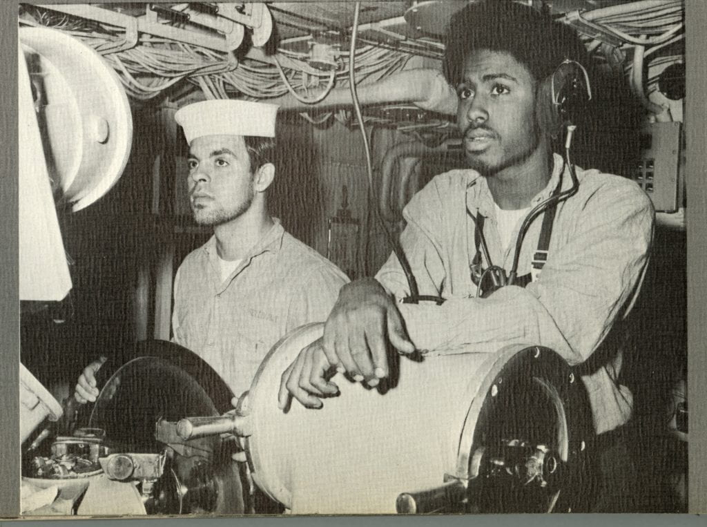 Black and white image of two men standing at helm, a light-skinned man grasps a wheel and a dark-skinned man wearing headphones rests his arms on a cylindrical drum