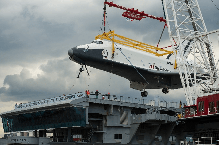 A large crane holds the space shuttle orbiter Enterprise, which looks like a bulky delta-winged airplane, above the flight deck of the former aircraft carrier Intrepid.