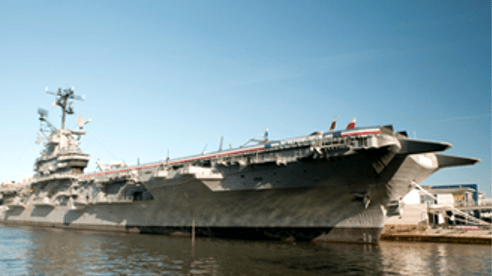 Sample image of the USS Intrepid docked on a body of water, a large gray floating military airfield that is as long as three football fields.