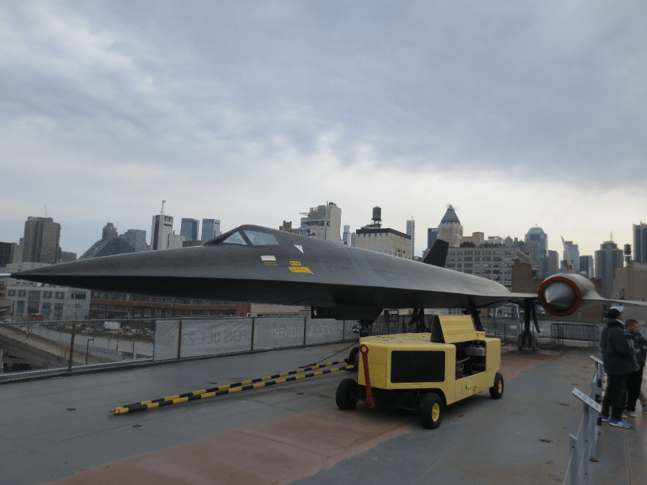 The black A-12 jet is in the center of the frame, facing left. It is long and narrow with a small cockpit on top and a large pointed jet engine on its thin delta wing. The NYC skyline is in the background.