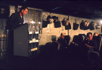 Color photo of a man standing at a podium addressing three men seated in chairs in front of him, helmets and bags hang from the wall in the background