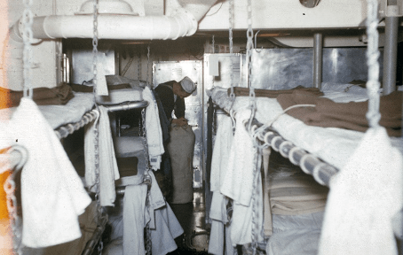 Color photo of berthing racks with unmade beds and towels hung around chains, a sailor in the background bends over a sea bag